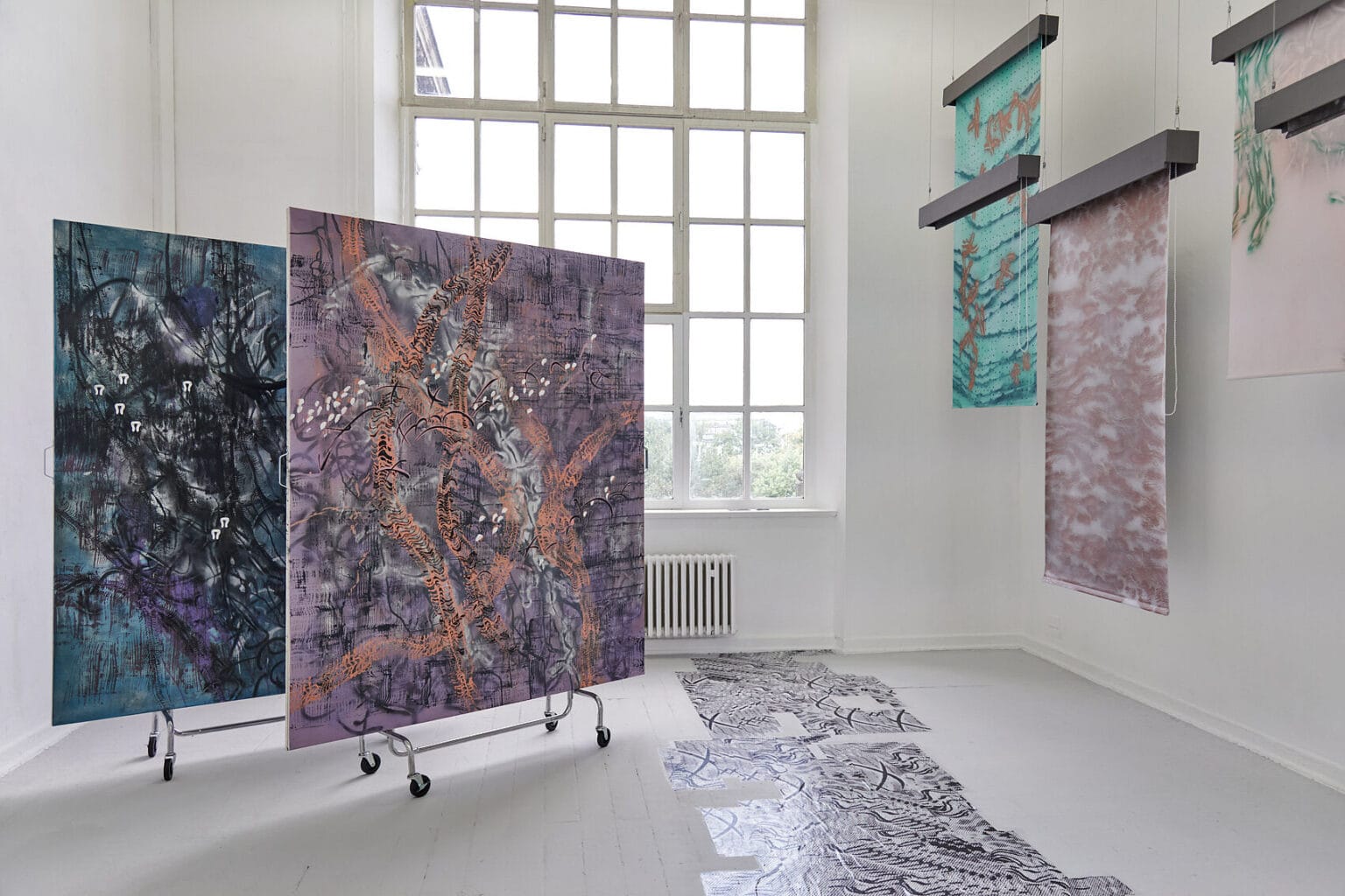 Foto: Ein Raum mit weißen Wänden, grauem Dielenboden und einem großen Fenster. Von der Decke hängen Fäden, an denen flache, herabhängenden Objekte befestigt sind. Auf dem Boden liegt eine Gruppe flacher, reflektierender, gemusterter Objekte. Links im Raum stehen zwei großformatige Leinwandarbeiten, die auf einem mit Rollen versehenen Gerüst stehen.