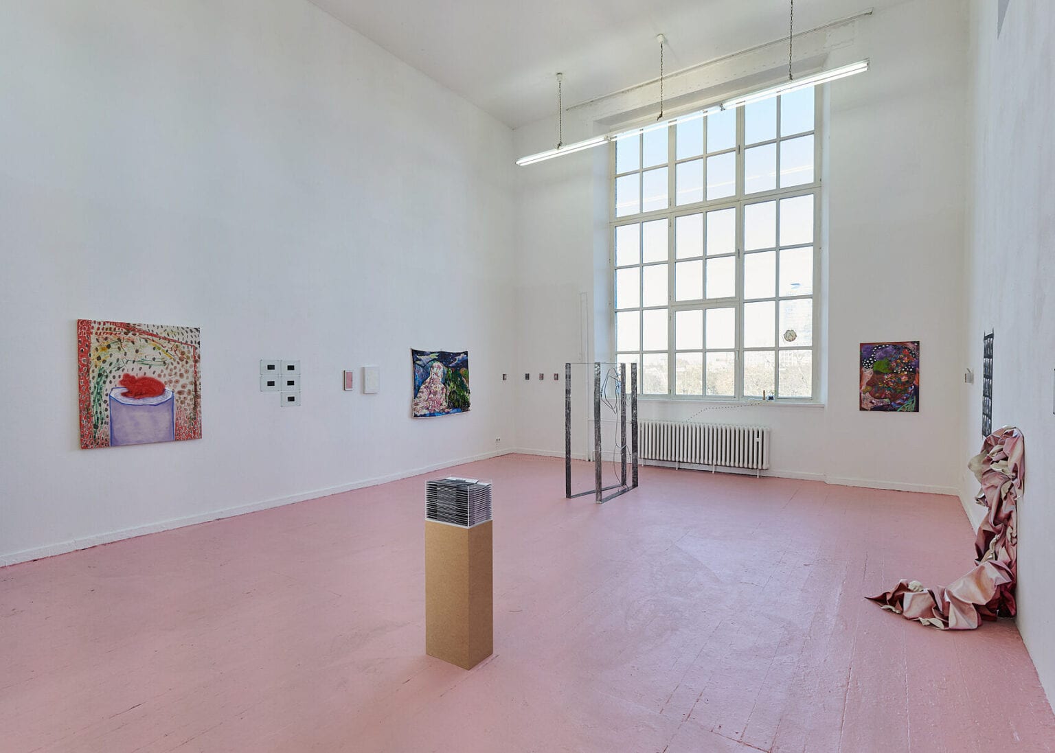 Foto: Ansicht eines Raumes mit weißen Wänden, rosanem Dielenboden und einem großen Fenster. An den Wänden befinden sich Malereien und flache Objekte in Klein- und Mittelformaten. Eines von ihnen reicht von der Wand herunter auf den Boden. Im Raum stehen zwei Skulpturen. Ein weiteres Objekt befindet sich auf einer der Fensterscheiben.