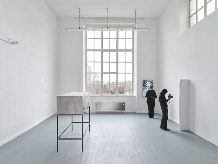 Foto: Ein Raum mit weißer Wand und grauem Boden mit einem großem Fenster und einem schmalen Fenster. An den Wänden befestigt ein Bildschirm und ein Objekt. Auch im Raum befindet sich ein Gegenstand. Eine Person mit Kamera steht in Nahsicht vor einem der Objekte. Eine weitere Person steht vor dem Bildschirm.