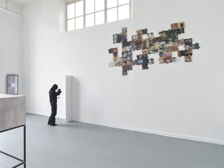 Foto: Ansicht eines Raumes mit weißer Wand und grauem Boden. Oben an der Wand ein schmales Fenster. An den Wänden befestigt ein Bildschirm und Objekte. Auch im Raum befindet sich ein Gegenstand. Eine Person mit Kamera steht nah vor einem der Objekte.