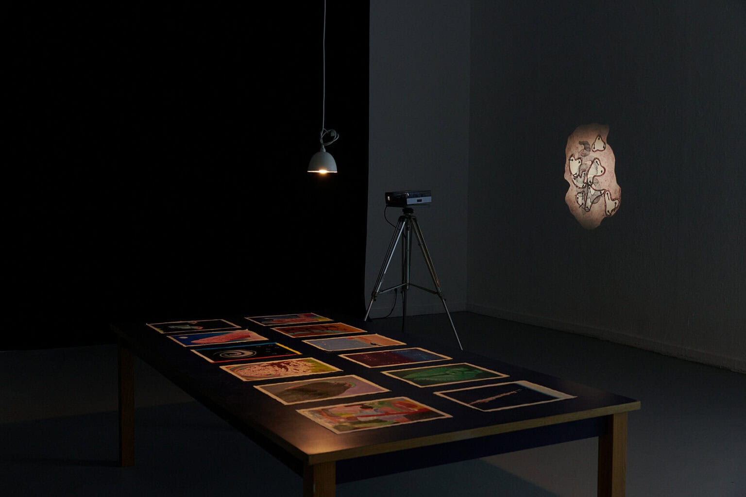Foto: Ansicht eines abgedunkelten Raumes mit weißen Wänden und dunklem Boden. Im Raum steht ein schwarzer Tisch, der von dem warmen Licht einer herabhängenden Deckenleuchte angestrahlt wird. Auf dem Tisch liegen in zwei Reihen Arbeiten auf Papier. Vor einer der Wände steht ein Projektor auf einem Stativ, der eine gemusterte Projektion an die Wand wirft.