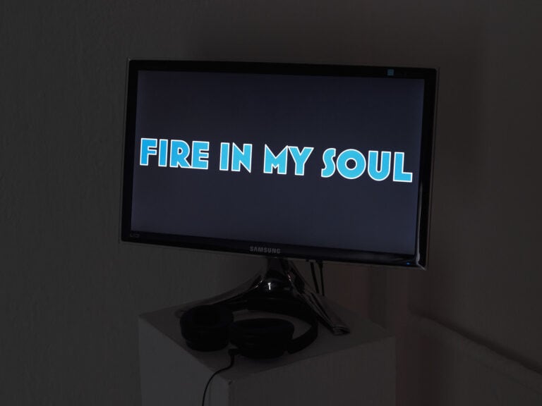 Foto: Nahaufnahme eines verkabelten Bildschirms auf einem Sockel in einem dunklen Raum. Auf dem Display zu sehen die Typografie "FIRE IN MY SOUL", in blauen Buchstaben mit weißer Rahmung.
