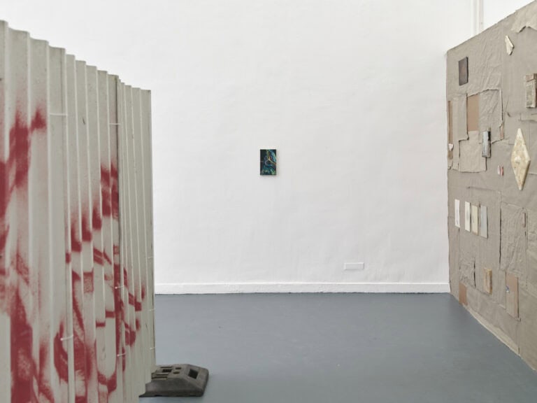 Foto: Ansicht eines Raumes mit weißer Wand und grauem Boden. Links eine Stellwand mit Betonfüßen, rechts eine Stellwand die mit Stoff behangen ist. Auf ihr befinden sich Zeichnungen und Objekte. An der weißen Wand hängt eine kleinformatige Malerei.