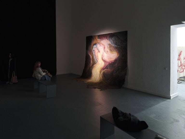 Foto: Ansicht eines abgedunkelten Raumes mit weißen Wänden und grauem Boden. Zwei Sitzbänke, auf der eine Person platz genommen hat. An der Wand lehnend befindet sich eine Malerei, die über Wand und Boden hängt. Sie wird von stellenweise von einem warmen Licht angestrahlt.