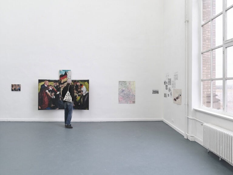 Foto: Ansicht eines Raumes mit weißen Wänden und grauem Boden, auf der rechten Seite ein Fenster. An der Wand befinden sich sich Arbeiten in Klein- und Mittelformaten. Vor einem der Arbeiten befindet sich ein Mensch, der die Arbeit in Nahsicht betrachtet.