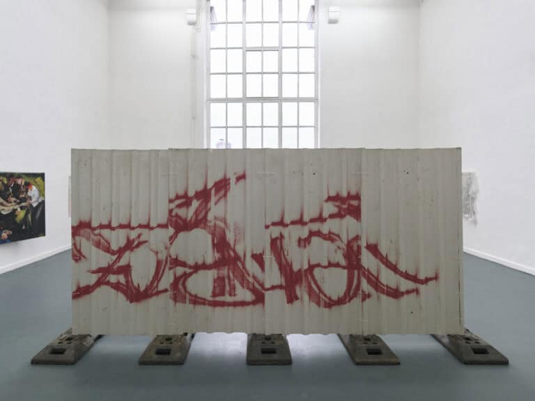 Foto: Ansicht eines weißen Raumes mit grauem Boden und großen Fenster. Im Vordergrund des Fotos Befindet sich eine große aufgestellte Wand mit Betonfüßen, die mit Farbe bearbeitet wurde. Im Hintergrund hängen zwei Arbeiten an der Wand.