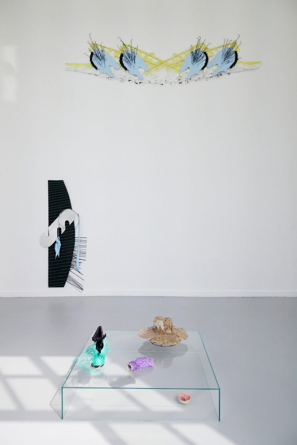 Foto: Ein Raum mit weißen Wänden, an denen auf unterschiedlicher Höhe Objekte hängen. Auf dem grauen Boden des Raumes eine Gruppe von Objekten aus unterschiedlichen Materialien.