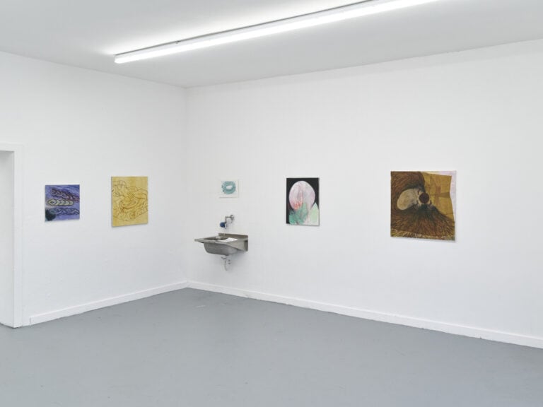 Foto: Ansicht eines Raumes mit weißen Wänden und grauem Boden. An den Wänden befinden sich Malereien und Papierarbeiten in Klein- und Mittelformaten und ein Waschbecken.