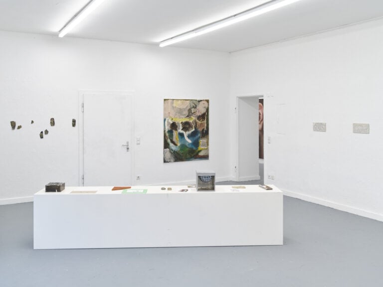 Foto: Ansicht eines Raumes mit weißen Wänden und grauem Boden. An den Wänden befinden sich Malereien in Klein- und Großformaten und Objekte. Im Raum steht ein heller, länglicher Sockel, auf dem Objekte und Papiere liegen.