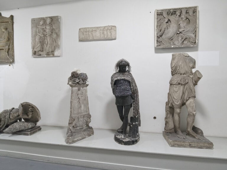 Foto: Raumansicht. Eine weiße Wand, davor ein länglicher, grauer Sockel. An der Wand befinden sich Reliefs und auf dem Sockel Skulpturen, die zum Teil mit Material behangen sind.