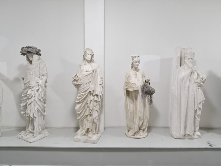 Foto: Ansicht eines Raumes mit weißer Wand und die Teilansicht eines länglichen, grauen Sockels. Auf dem Sockel befinden sich vier helle Skulpturen, die zum Teil mit Material behangen sind.