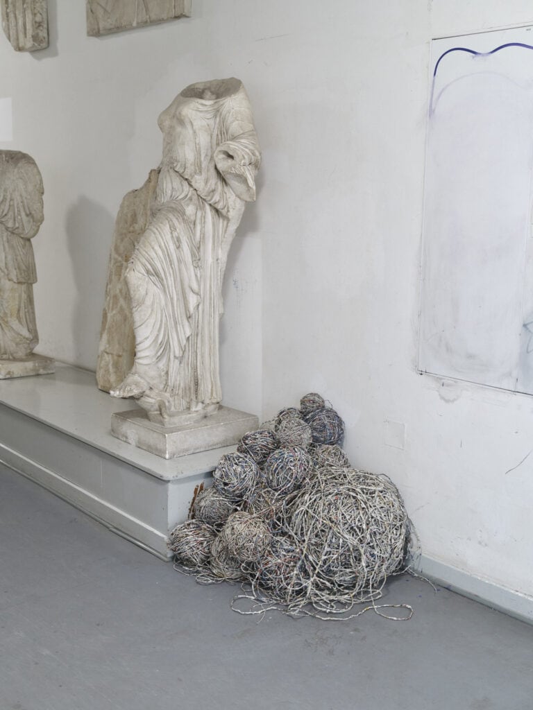 Foto: Nahaufnahme eines Raumes mit grauem Boden. In dem Raum ein hellgrauer länglicher Sockel, der aus dem Bildraum hinaus ragt. Auf dem Boden neben dem Sockel befinden sich zu verschiedenen Größen zusammengerollte Kneule aus Materialfäden. Auf dem Sockel befinden sich helle Skulpturen.