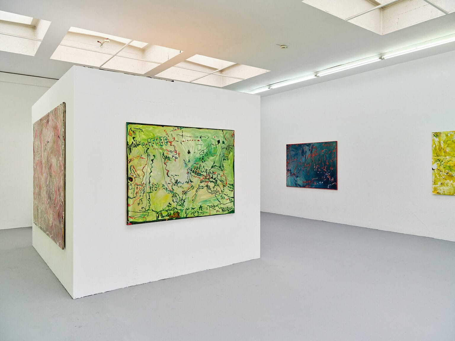 Foto: Ansicht eines Raumes mit weißen Wänden, grauem Boden und Deckenfenstern. Im Raum befindet sich ein großer weißer Kubus, auf dem mittel- und großformatige Malereien hängen. An den Wänden hängen ebenfalls Malereien.