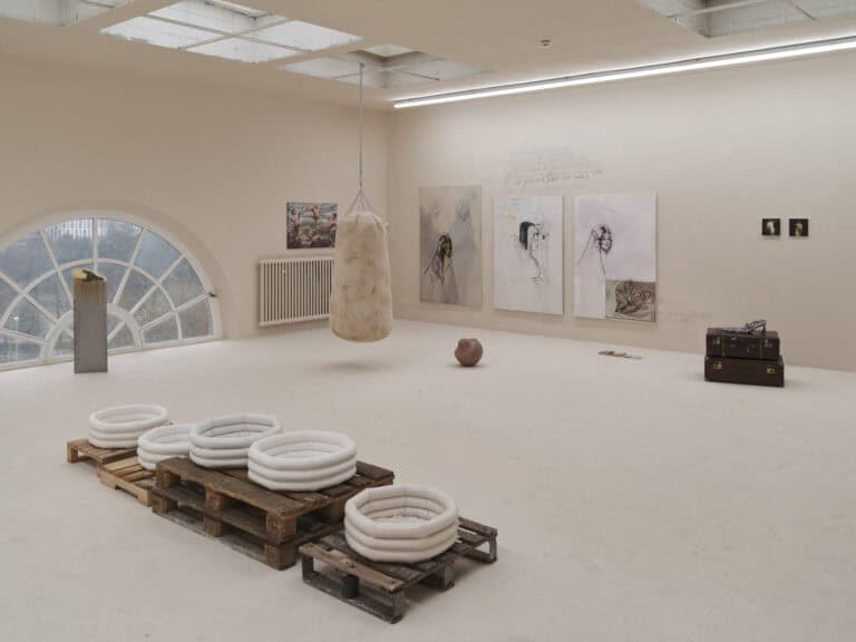 Foto: Ansicht eines Raumes, dessen Decke, Wände und Boden in einem hellen Cremeton gestrichen sind. Ein Bogenfenster in Form eines Halbkreises. Auf der Boden befinden sich mehrere Skulpturen, einige sind auf Paletten gestapelt, andere hängen von der Decke. An den Wänden befinden sich mehrere klein- bis großformatige Bilder. An der Wand ist zum Teil verwischte Schrift aufgebracht.