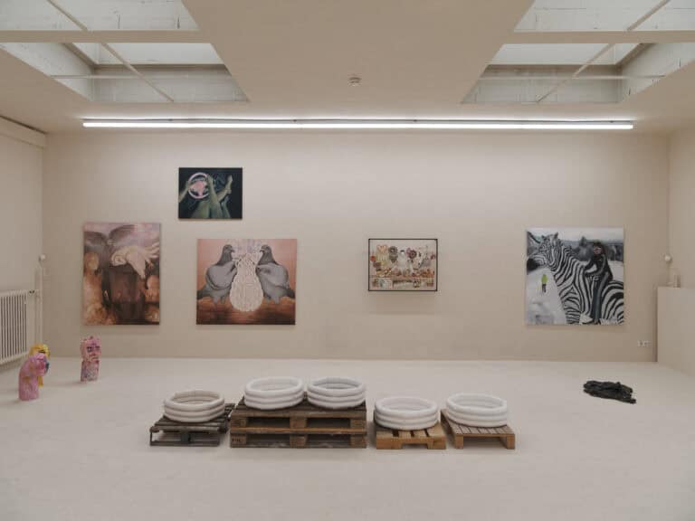 Foto: Ansicht eines Raumes, dessen Decke, Wände und Boden in einem hellen Cremeton gestrichen sind. Auf der Boden befinden sich mehrere Skulpturen, einige sind auf Paletten gestapelt. An den Wänden befinden sich fünf mittel- bis großformatige Bilder.