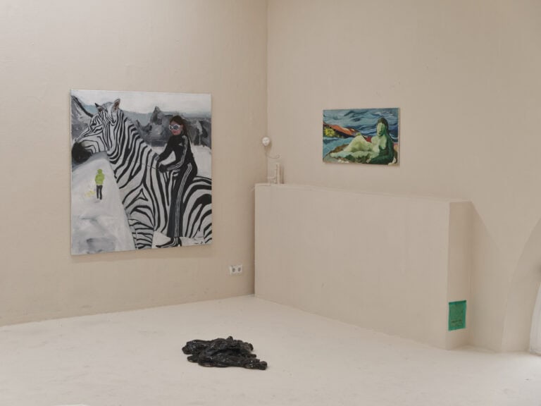 Foto: Ansicht der Ecke eines Raumes. Wände und Boden sind in einem hellen Cremeton gestrichen. Auf der Boden befindet sich ein dunkles, reflektierendes Objekt. An der linken Wand befindet sich eine großformatige Leinwand. Genau in der Ecke des Raumes eine kleine, skulpturale Arbeit. An der rechten Wand hängt eine mittelformatige Leinwand Arbeit und bodennah ein grünes Rechteck.