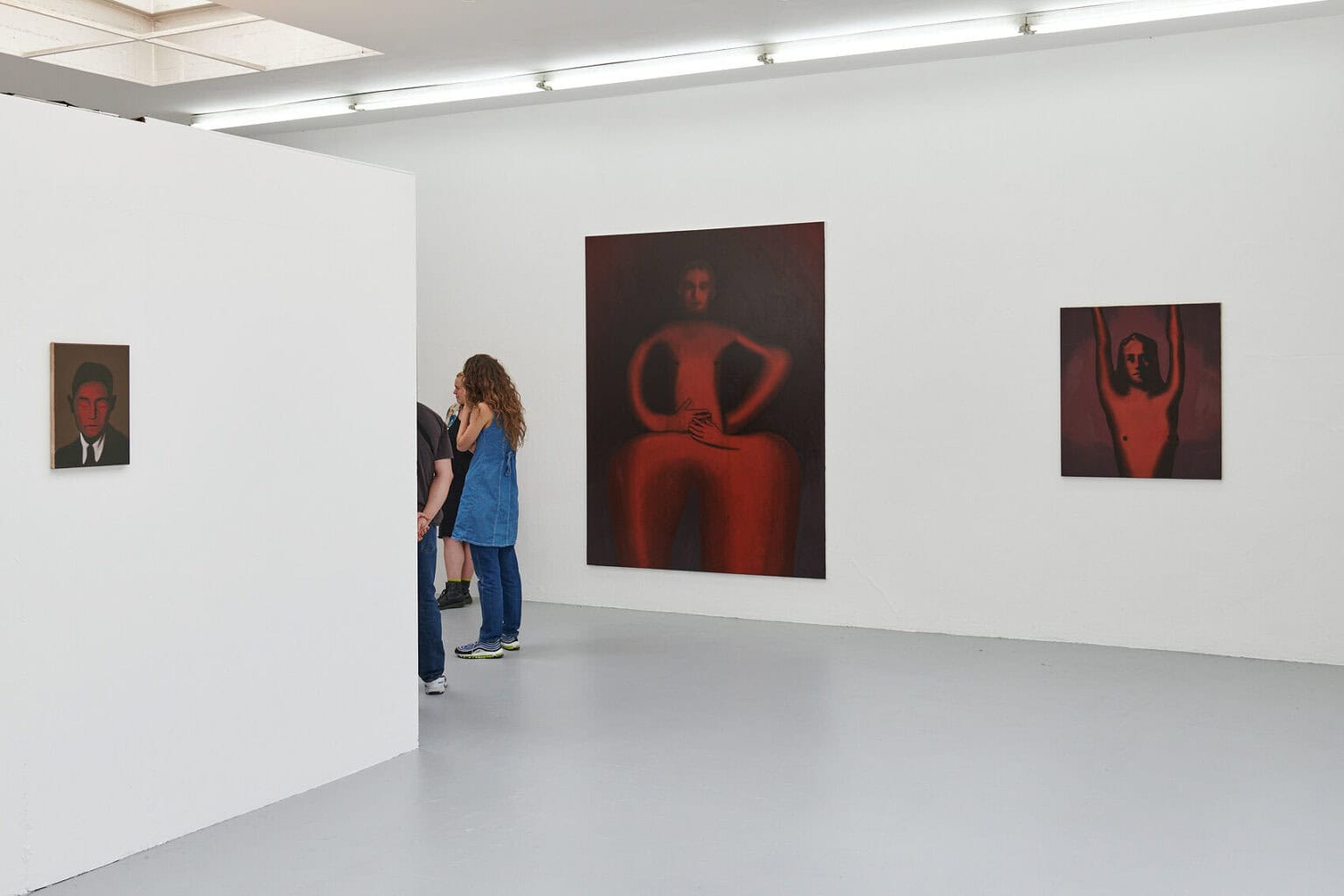 Foto: Ein Raum mit weißen Wänden und grauem Boden. Im Raum stehen Menschen. An den Wänden hängen Leinwandarbeiten im Klein-, Mittel- und Großformat.