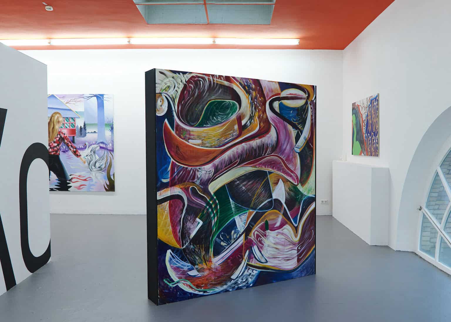 Foto: Ansicht eines Raumes mit weißen Wänden, grauem Boden und einem kleinen Bogenfenster. Die Decke ist rot gestrichen. An den Wänden hängen Malereien im Mittel- und Großformat. Im Raum stehen flache, ebenfalls mit Farbe bearbeitete Objekte.