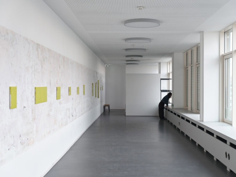 Foto: Ansicht eines weißen Raumes mit dunklem Boden. An der linken, teils unebenen Wand, befindet sich elf Leinwände in Klein- bis Mittelformaten. Hinten im Raum stehen Hocker. Rechts ist eine Fensterfront mit teilweise herabgesenkten Rollos zu sehen. An der Fensterbank ein nach Vorn übergebeugter Mensch.