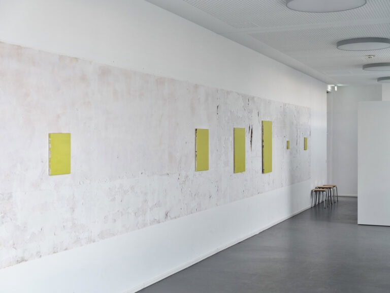 Foto: Ansicht eines weißen Raumes mit dunklem Boden. An der linken, teils unebenen Wand befinden sich sechs klein- bis mittelformatige Leinwände. Hinten im Raum stehen mehrere Hocker.