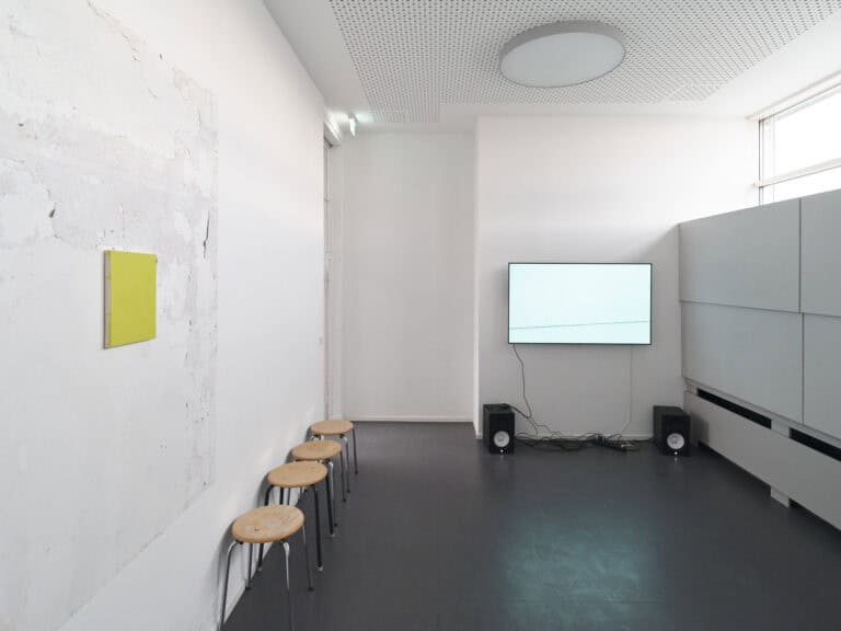 Foto: Ansicht eines weißen Raumes mit dunklem Boden. An der linken, teils unebenen Wand befindet sich eine kleinformatige Leinwand. Unten an der Wand stehen vier Hocker. An einer anderen Wand hängt ein Bildschirm mit zwei Boxen. Die Fensterfront ist teilweise abgedeckt.