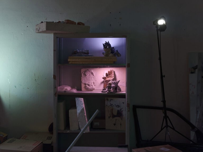 Foto: Nahaufnahme eines Regals in einem halbdunklen, mit vielen Gegenständen gefüllten Raum. Ein Regal in und auf dem Dinge Lagern. In zweien der Regalsegmente sind Lichter angebracht, die den Regalinhalt beleuchten. Rechts neben dem Regal ein eingeschaltetes Standlicht.