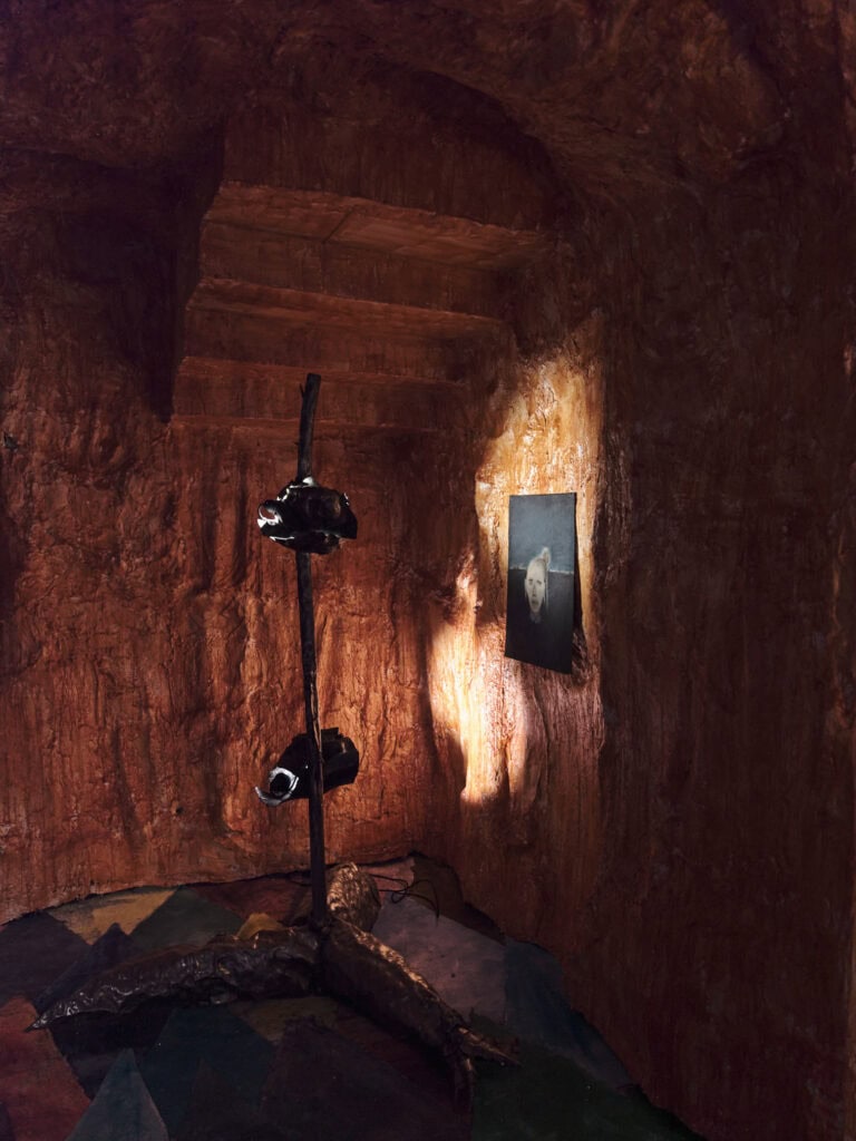 Foto: Ansicht eines Raumes mit rot-braunen Wänden und Decke. Eine stativartige Konstruktion mit zwei Scheinwerfern und einem großen Stativfuß vor der unebenen Wand. Die Lichter werfen zwei helle Lichtkegel an die Wand. An der Wand ist eine Malerei angebracht, zum Teil angestrahlt wird. Der Boden ist mit bunten Rautenformen bemalt.