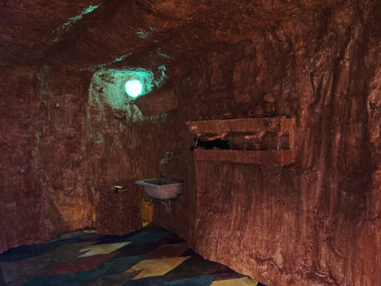 Foto: Ansicht eines Raumes mit unebenen, rot-braun bemalten Wänden und Decke. In die Wand sind rot-braun bemalte Gegenstände und ein Waschbecken eingelassen. Der Boden ist mit bunten Rautenformen bemalt. In der Ecke leuchtet ein grünes, rundes Licht, das ebenfalls in die Wand eingelassen ist. Das Licht wird von der Wand reflektiert.