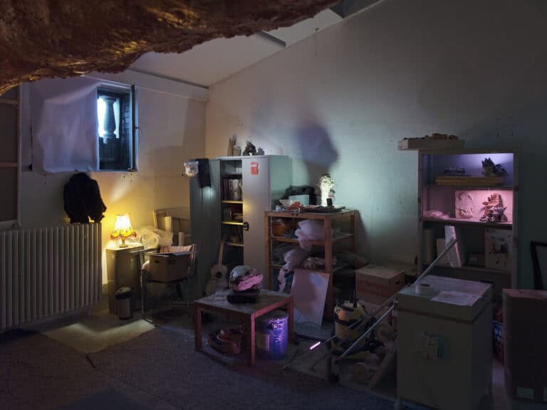 Foto: Ansicht eines halbdunklen, mit vielen Gegenständen gefüllter Raumes mit kleinem Fenster und weißen Wänden. In, auf und unter Möbeln gestapelte Gegenstände. Eine gelb leuchtende Lampe auf einem Karton. Weitere Lichtquellen in Regalen.