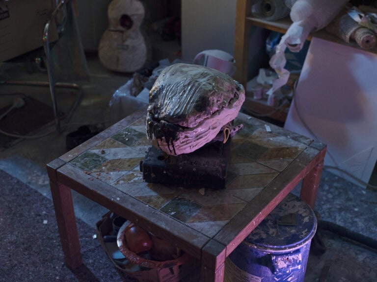 Foto: Nahaufnahme eines kleinen, gekachelten, quadratischer Tisches in einem mit vielen Gegenständen gefüllten Raum. Auf dem Tisch ein glänzender, fester Gegenstand. Der Tisch und das Objekt sind in rötliches Licht getaucht.