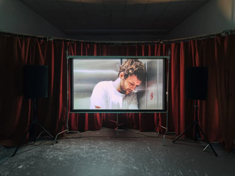 Foto: Ansicht eines abgedunkelten Raumes mit grauem Boden. Zentral im Foto ein Bildschirm vor einer Wand aus roten Vorhängen. Daneben zwei Lautsprecher auf Stativen.