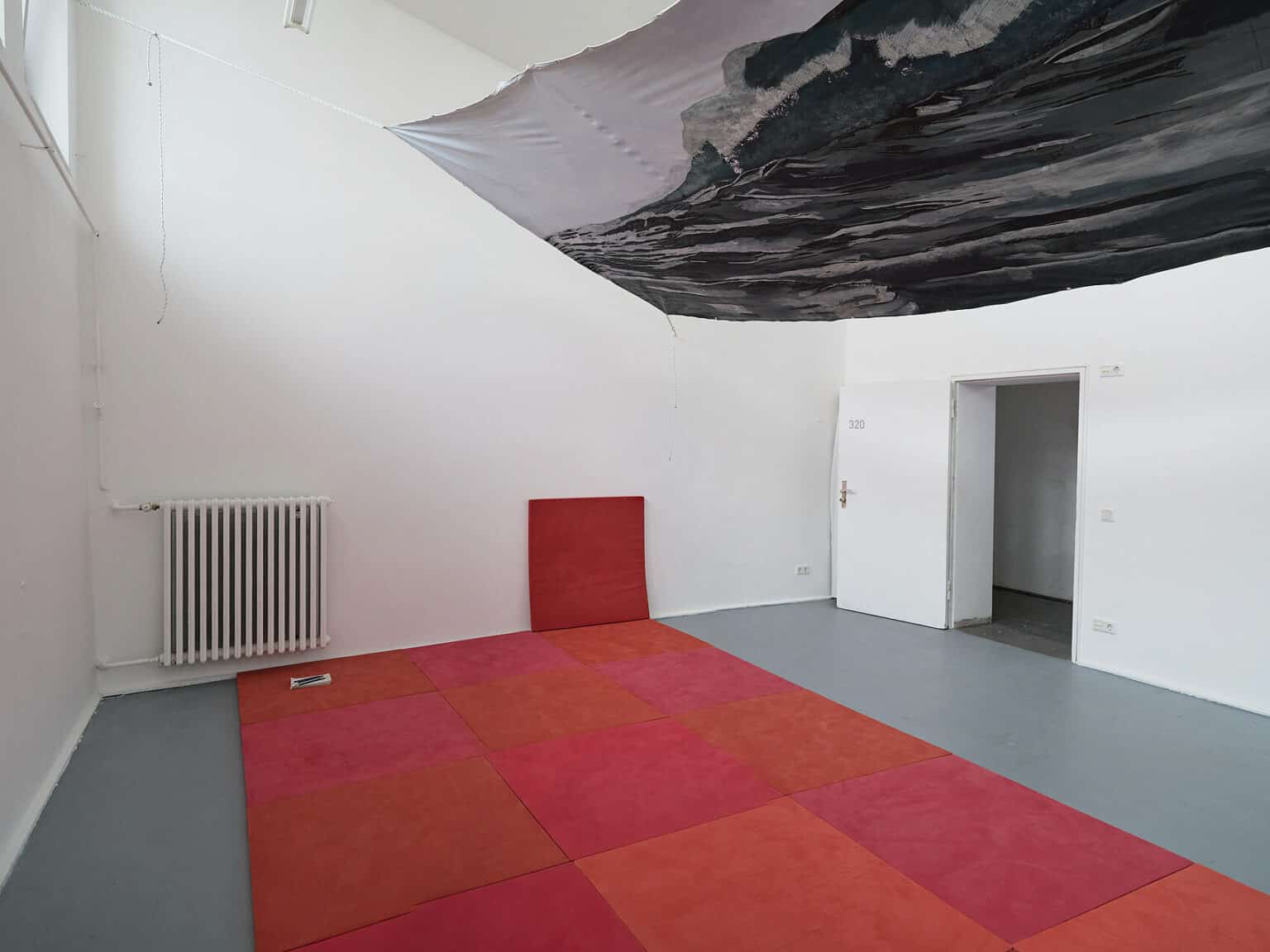 Foto: Ein Raum mit weißen Wänden und grauem Boden. Deckennah ist ein Textil horizontal im Raum an Seilen aufgespannt. Der Boden ist mit quadratischen Objekten in verschiedenen rot- und orangenen ausgelegt. An einer der Wände lehnt eines der quadratischen Objekte. Die Tür ist geöffnet.