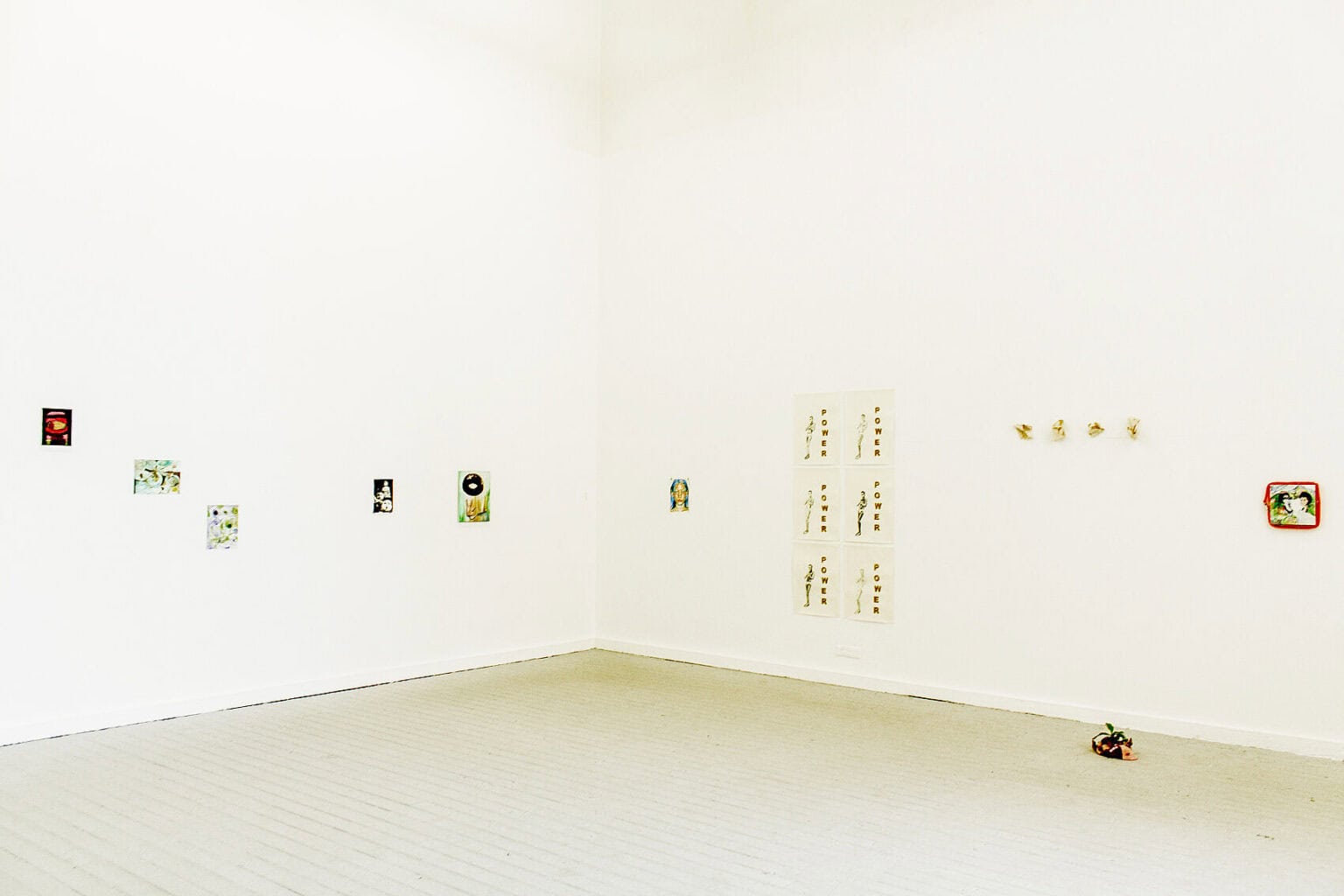 Foto: Die Ecke eines Raumes mit weißen Wänden und grauem Dielenboden. An den Wänden hängen, auf unterschiedlicher Höhe aber bodennah, kleinformatige Objekte. Auf dem Boden liegt ebenfalls ein kleines Objekt.