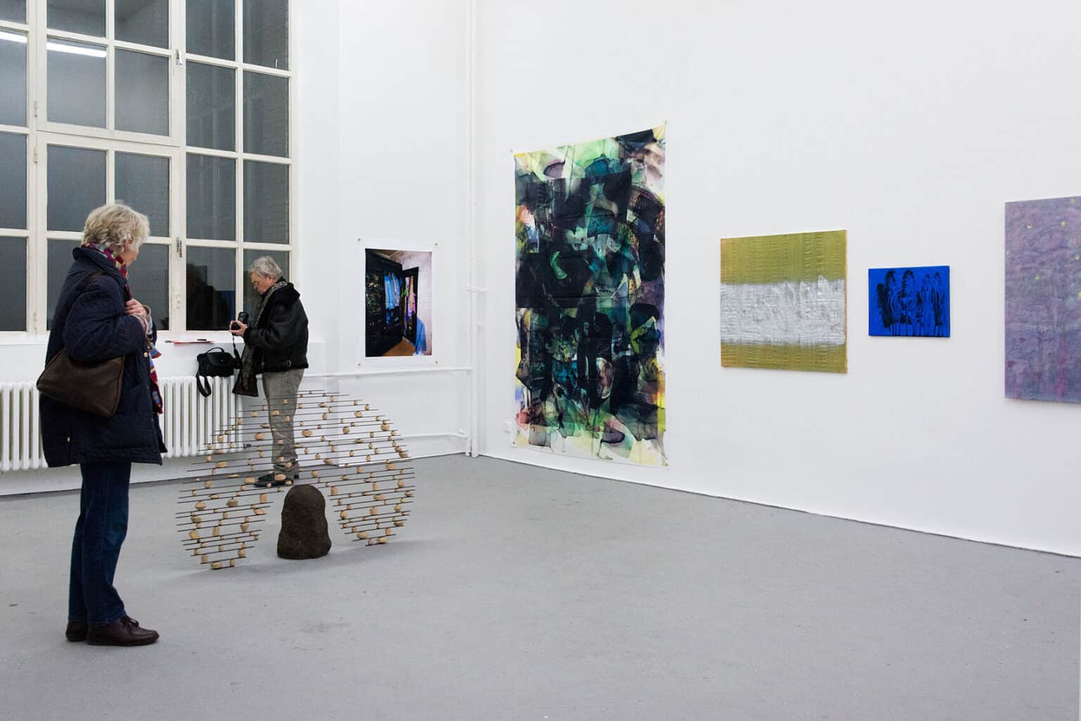 Foto: Ein Raum mit weißen Wänden, grauem Boden und einem großen Fenster. An der Wand befinden sich mehrere flache Objekte im Mittel- und Großformat. Im Raum steht eine Skulptur auf dem Boden. Im Raum befinden sich zwei Menschen.