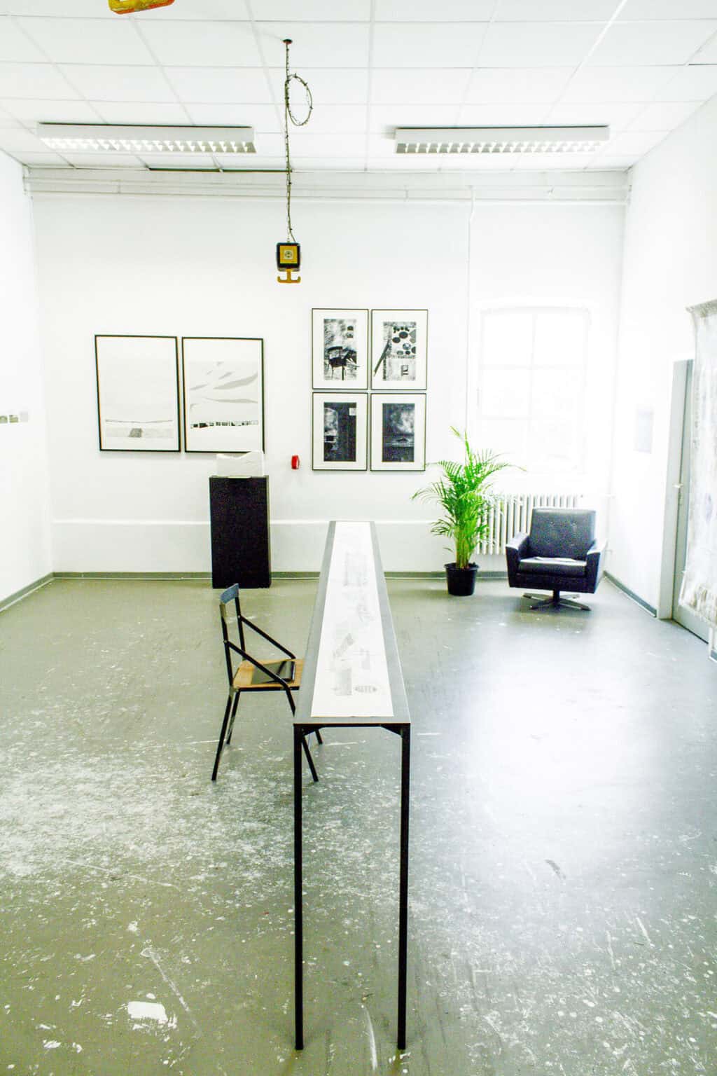 Foto: Ein Raum mit weißen Wänden und grauem, mit Farbflecken versehenem Boden. An der Wand hängen mehrere gerahmte Objekte. Von der abgehangenen Decke hängt eine Steckdosenleiste. Im Raum steht ein Sessel, eine Pflanze, ein Sockel mit einem Objekt, ein langer, schmaler Tisch auf dem ein langes Papier liegt und ein Stuhl.