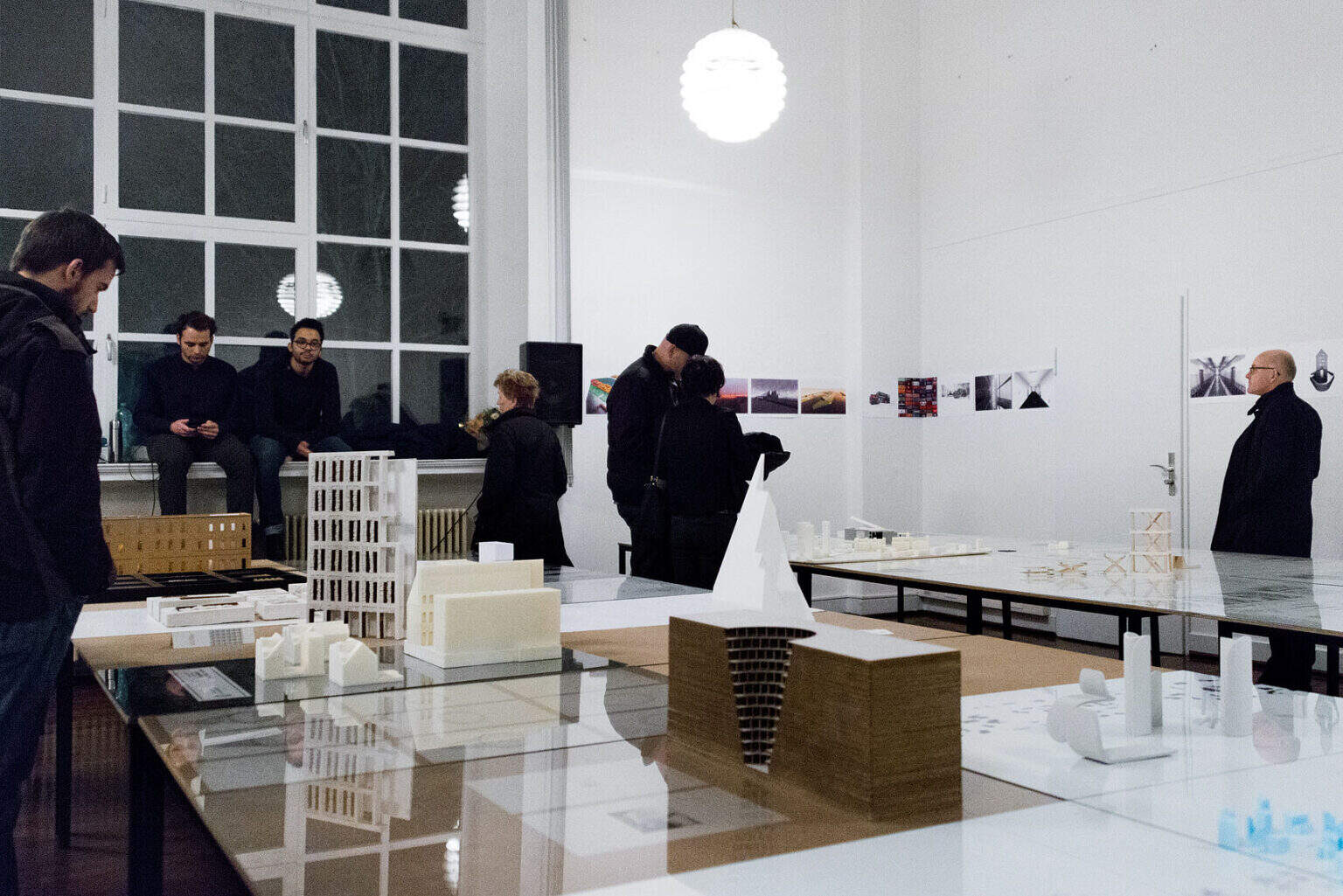 Foto: Ein Raum mit weißen Wänden und einem großen Fenster. An der Wand befinden sich bedruckte Papiere, die auf gleicher Unterkante in einem Streifen hängen, der sich über mehrere Wände erstreckt. Im Raum stehen Tische. Einige von ihnen sind mit Glasplatten bedeckt. Under den Glasplatten liegen Papiere, Fotografien, Zeichnungen usw. Auf den Glasplatten und Tischen stehen modellartige Objekte. Im Raum befinden sich Menschen.