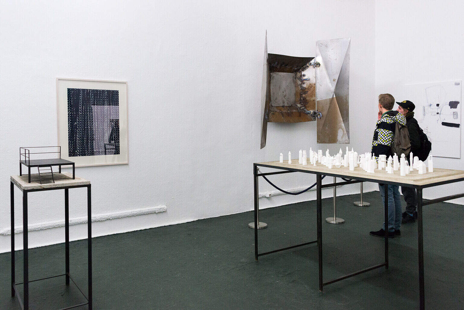 Foto: Ein Raum mit weißen Wänden und schwarzem Boden. An den Wänden befinden sich flache und plastische Objekte. Im Raum stehen Tische und Sockel, auf denen sich Objekte befinden. Durch den Raum bewegen sich Menschen.