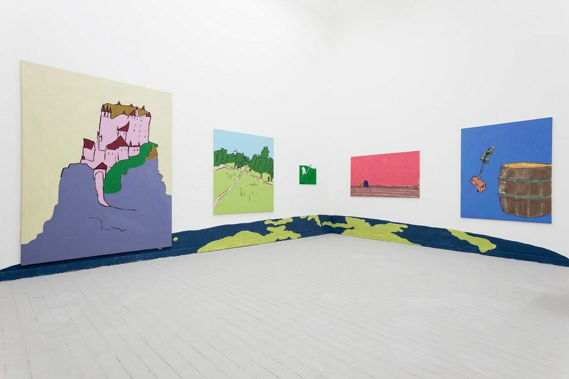 Foto: Ein Raum mit weißen Wänden und grauem Dielenboden. Die Fußleiste und Teile der darüberliegende Wand sind in der Form eines länglichen Halbkreises mit blauer und roter Farbe bemalt. An den Wänden hängen, auf ähnlicher Höhe, mehrere Malereien im Klein-, Mittel- und Großformat.