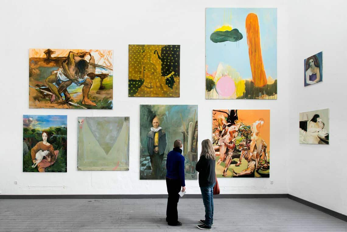 Foto: Ein Raum mit weißen Wänden und grauem Dielenboden. An den Wänden hängen mehrere Leinwände in unterschiedlichen Formaten neben- und untereinander.