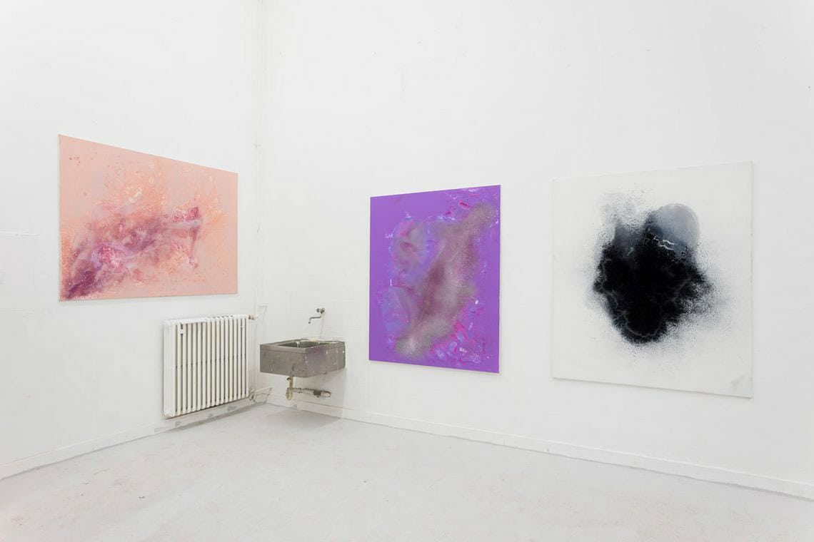 Eine Raumecke mit weißen Wänden und hellgrauem Boden. An der Wand ein Waschbecken und drei Leinwände in ähnlichem Format auf ähnlicher Höhe.