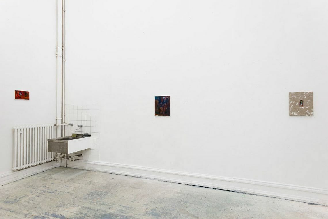 Foto: Ein Raum mit weißen Wänden und gräulichem Boden. An der Wand ein Waschbecken und drei kleinformatige Arbeiten auf Leinwand auf ähnlicher Höhe.