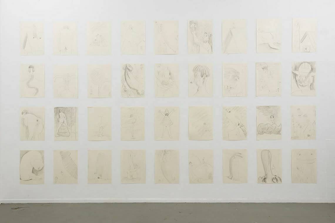 Foto: Eine weiße Wand und grauer Boden. An der Wand befinden sich, in vier Reihen hängend, Papierarbeiten im gleichem Format. Sie hängen im gleichen Abstand über- und untereinander.