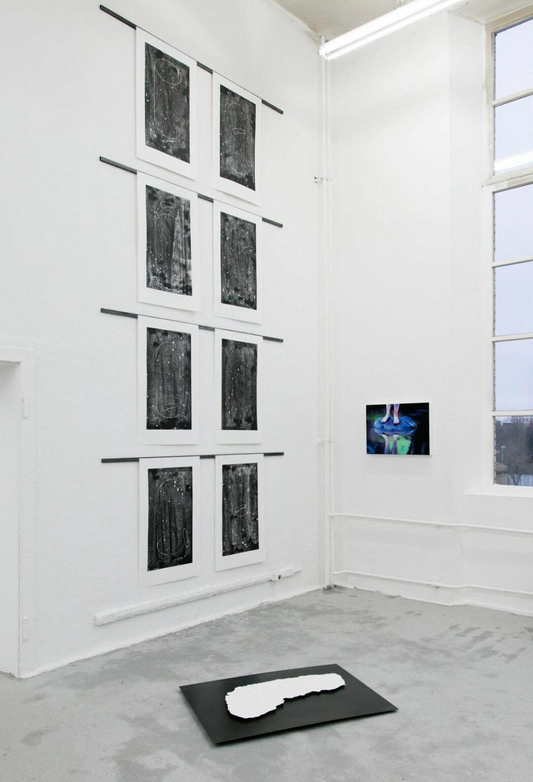 Foto: Ein Raum mit weißen Wänden, grauem Boden und einem großen Fenster. Über die gesamte Höhe der Wand sind im immer gleichen Abstand zueinander Leisten montiert, von denen großformatige, bedruckte Papiere herabhängen. Die Papiere hängen in Zweierreihen neben und in vier Reihen untereinander. An einer weiteren Wand hängt ein flaches Objekt. Auf dem Boden liegt ein weiteres flaches Objekt.