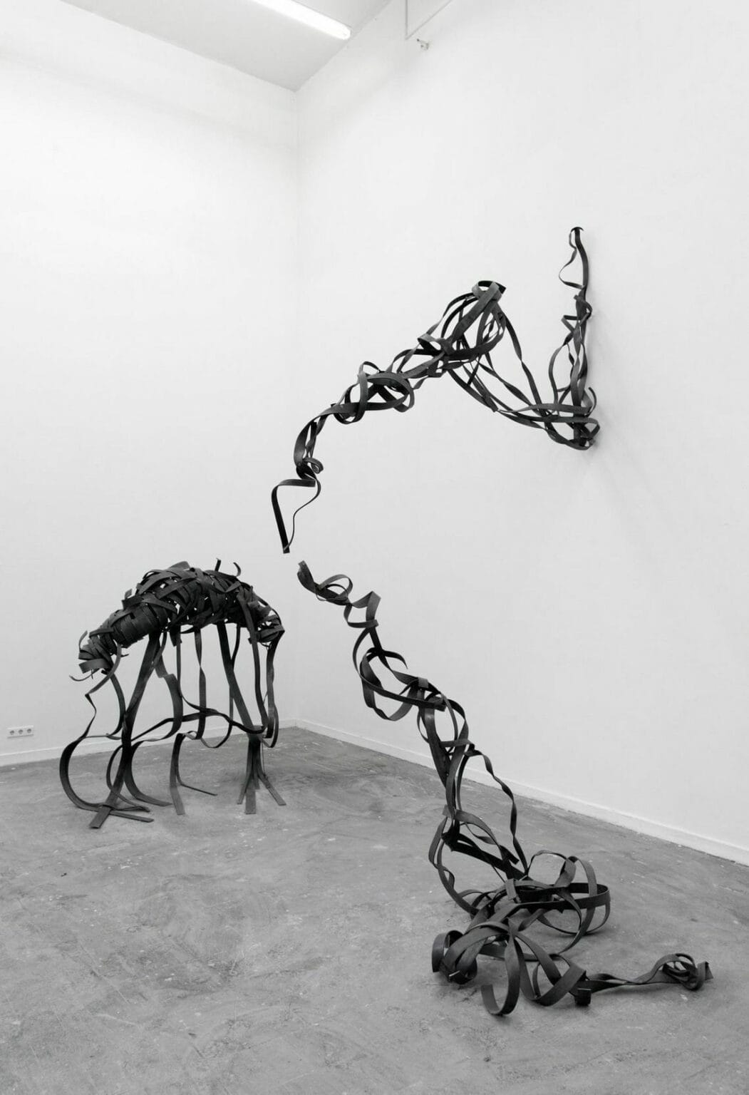 Foto: Ein Raum mit weißen Wänden und grauem Boden. Auf dem Boden des Raumes und an der Wand ist eine Gruppe von Skulpturen installiert, die aus dem gleichen Material zu bestehen scheint. Die Skulpturen kommen sich teilweise sehr nach, berühren sich aber nicht.