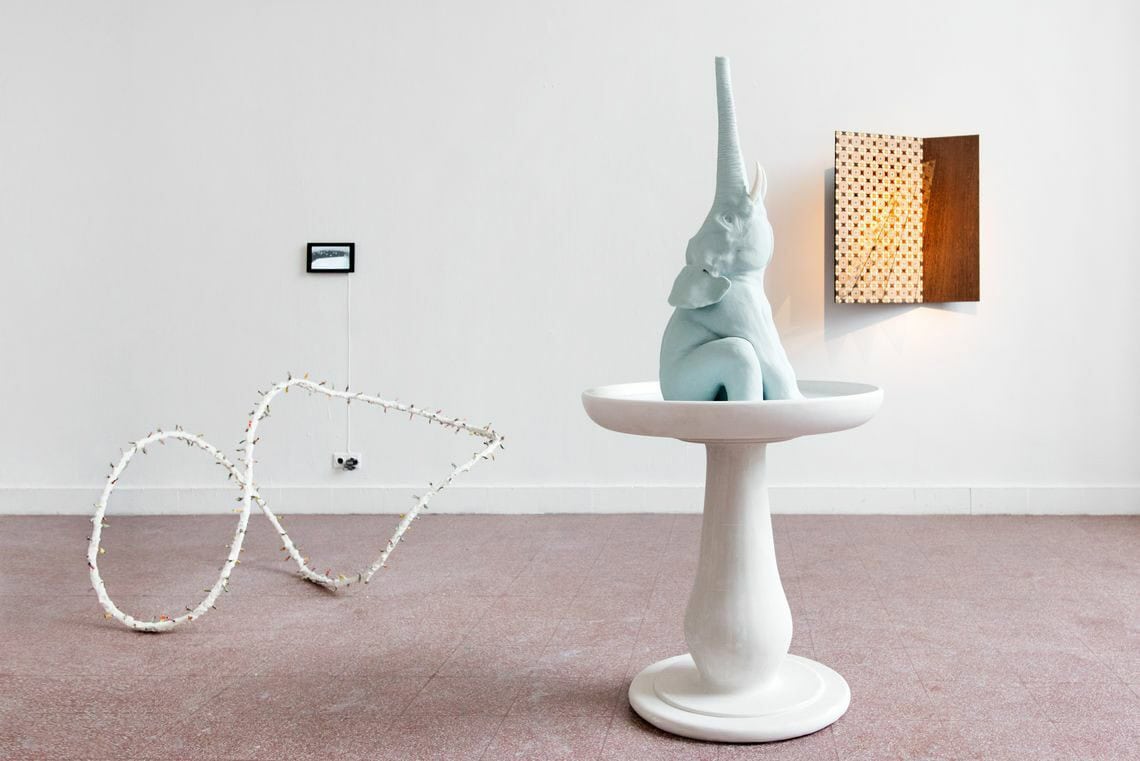 Foto: Ein Raum mit weißen Wänden und rotem Steinboden. Im Raum und an der Wand befinden sich mehrere Objekte aus unterschiedlichen Materialien.