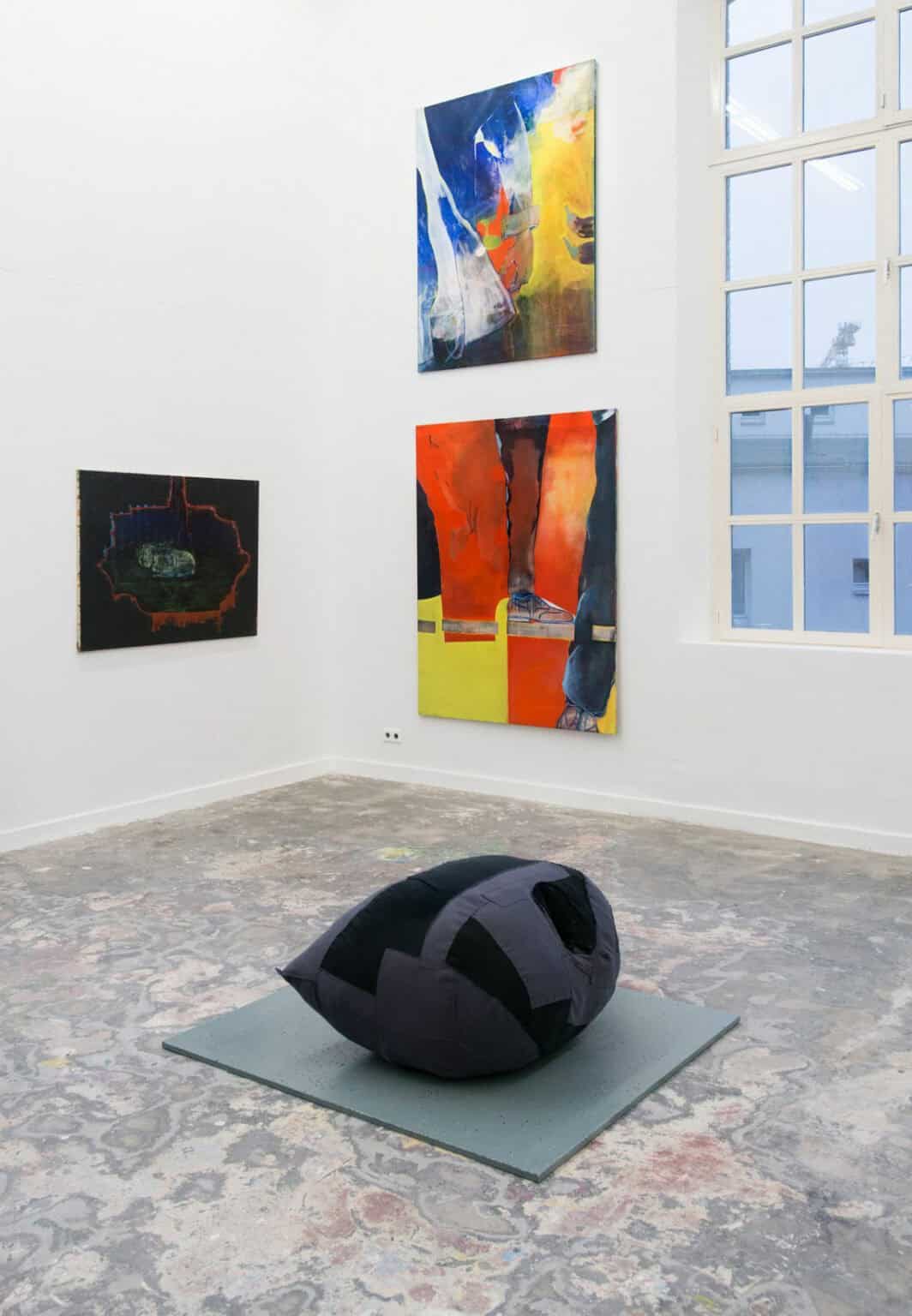 Foto: Ein Raum mit weißen Wänden, mehrfarbigem Boden in aschigen Tönen und einem großen Fenster. An der Wand hängen Leinwände in Groß- und Mittelformaten auf unterschiedlicher Höhe. Auf dem Boden des Raumes liegt eine Skulptur auf einem flachen Sockel.