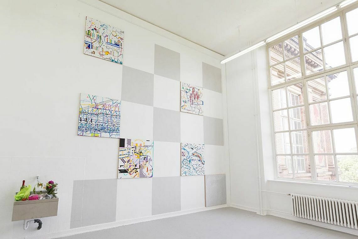 Foto: Ein Raum mit weißen Wänden, grauem Boden, einem großen Fenster und einem Waschbecken, in dem sich Blumen befinden. Auf die Wand sind, in der Musterung eines Schachbretts, graue auf die Wand aufgemalte Quadrate und quadratische Leinwandarbeiten angebracht.