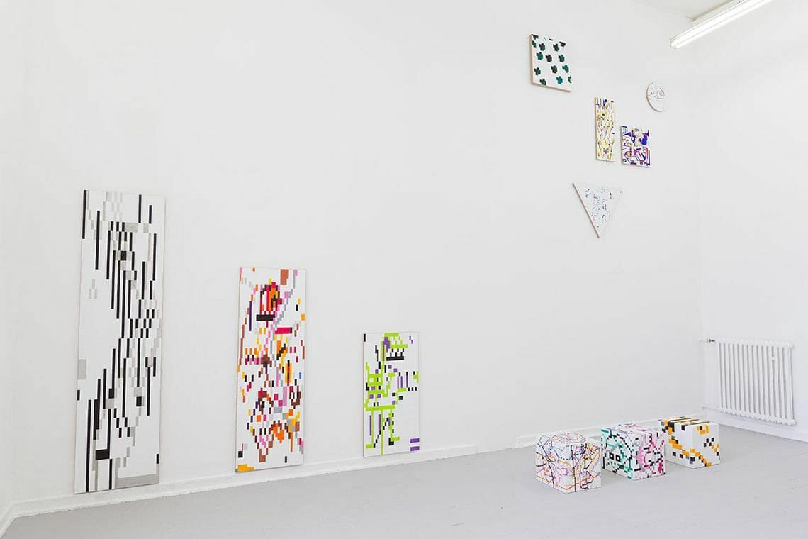 Foto: Ein Raum mit weißen Wänden und grauem Boden. An der Wand hängen, auf unterschiedlichen Höhen Leinwandarbeiten. Manche von ihnen hängen bodennah, andere hochoben an der Wand. Die Leinwände sind rechteckig, rund und dreieckig. Im Raum stehen drei quadratische Skulpturen in einer Reihe nebeneinander.