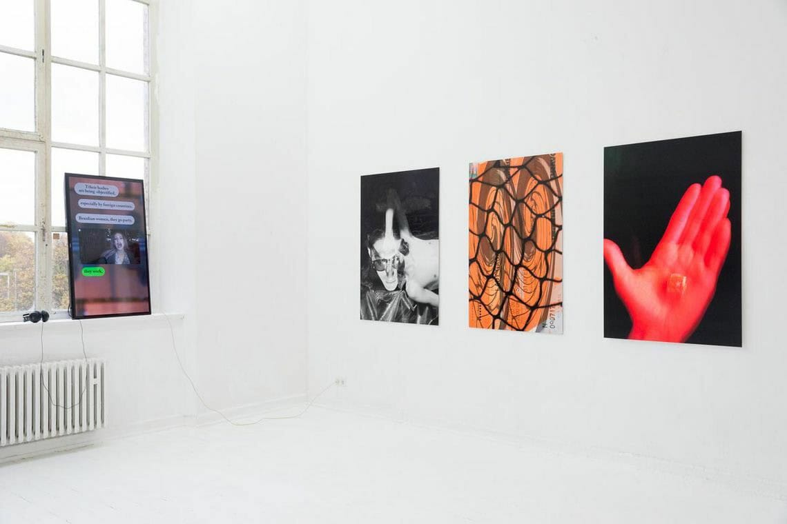 Foto: Ein Raum mit weißen Wänden, weißem Boden und einem Fenster. Auf der Fensterbank lehnt ein eingeschalteter Bildschirm in Hochformat, der mit Kopfhörern verbunden ist. An der Wand befinden sich drei flache Objekte, in gleichem Format, auf gleicher Höhe und im gleichen Abstand zueinander hängend.