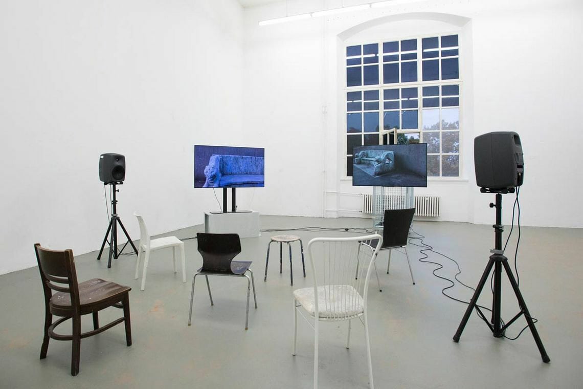 Foto: Ein Raum mit weißen Wänden, grauem Boden und einem großen Bogenfenster. Im Raum verteilt stehen zwei Bildschirme und zwei Lautsprecher auf Stativen und mehre Stühle und Hocker, die allesamt unterschiedlich aussehen.
